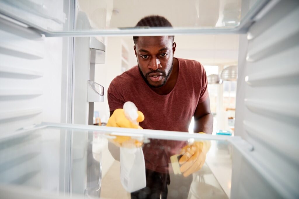 Man cleaning the interior of a refrigerator with cleaning gloves, a sponge, and a cleaning spray bottle