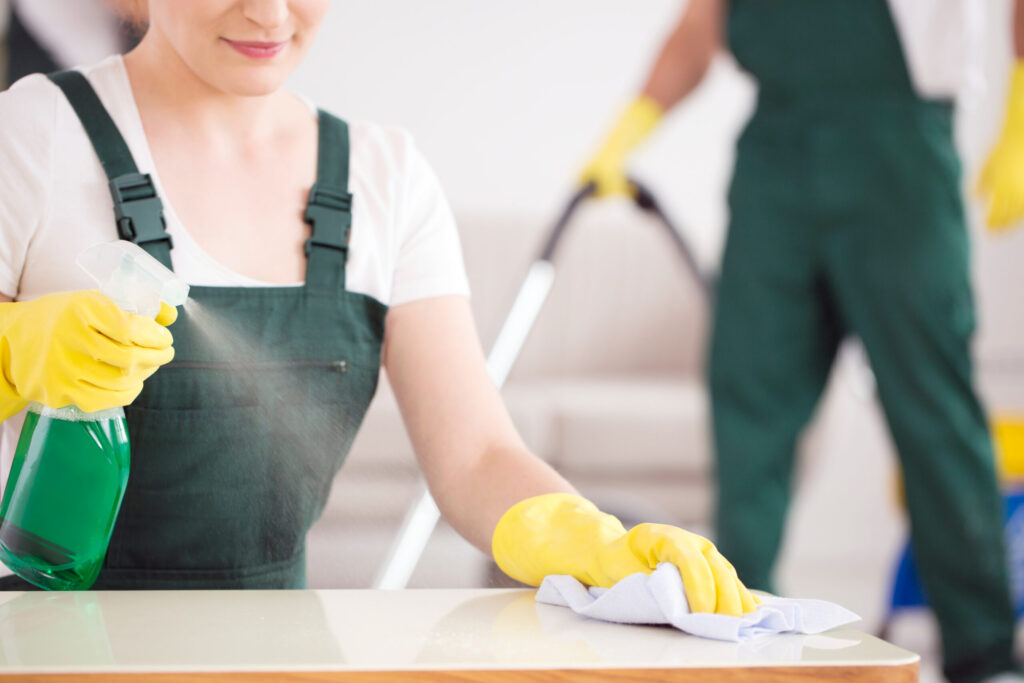 Female custodian cleaning a table with yellow rubber gloves on and using a green cleaning solution and cloth