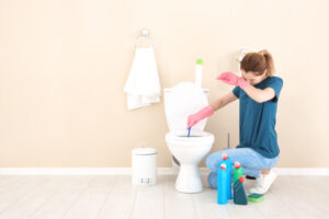 Woman kneeling down and cleaning a toilet while holding her hand to her face indicating the chemical smell is strong