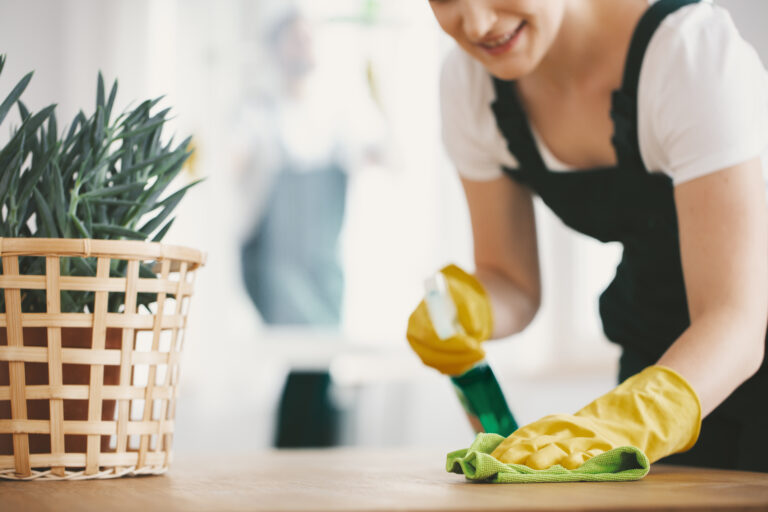 Woman cleaning a wooden table with yellow rubber gloves on, a cloth, and a cleaning solution