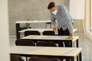Male teacher with a face mask on cleaning desks and chairs with a cleaning solution and cloth