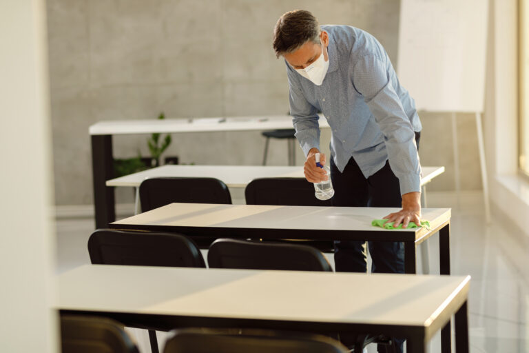 Male teacher with a face mask on cleaning desks and chairs with a cleaning solution and cloth
