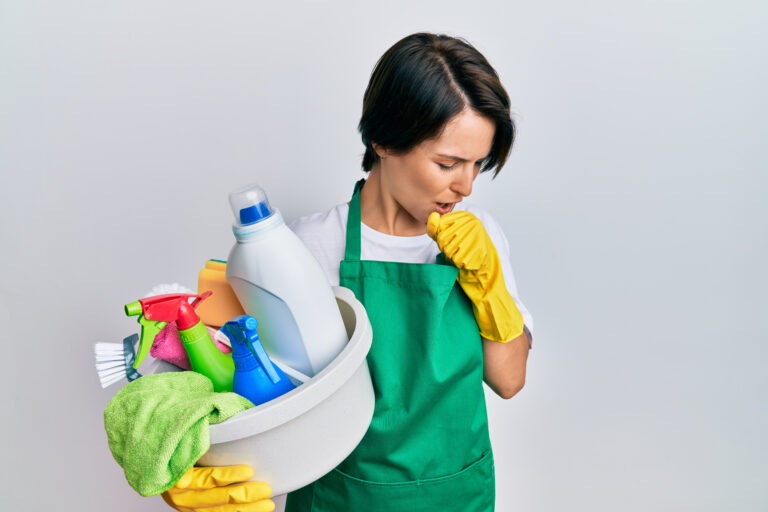 Young woman coughing while holding a bucket of cleaning supplies indicating a harsh chemical smell