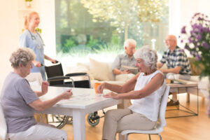 Two senior woman working on a puzzle together at a table while two older men have a conversation in the background at a senior living facility